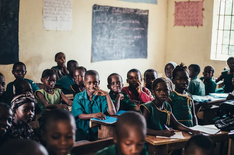 A full classroom of Rwandan children at wooden desks looking toward the blackboard